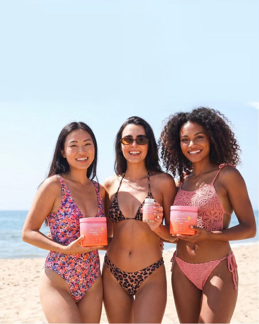 Three women on a beach holding colorful drinks against a clear blue sky.
