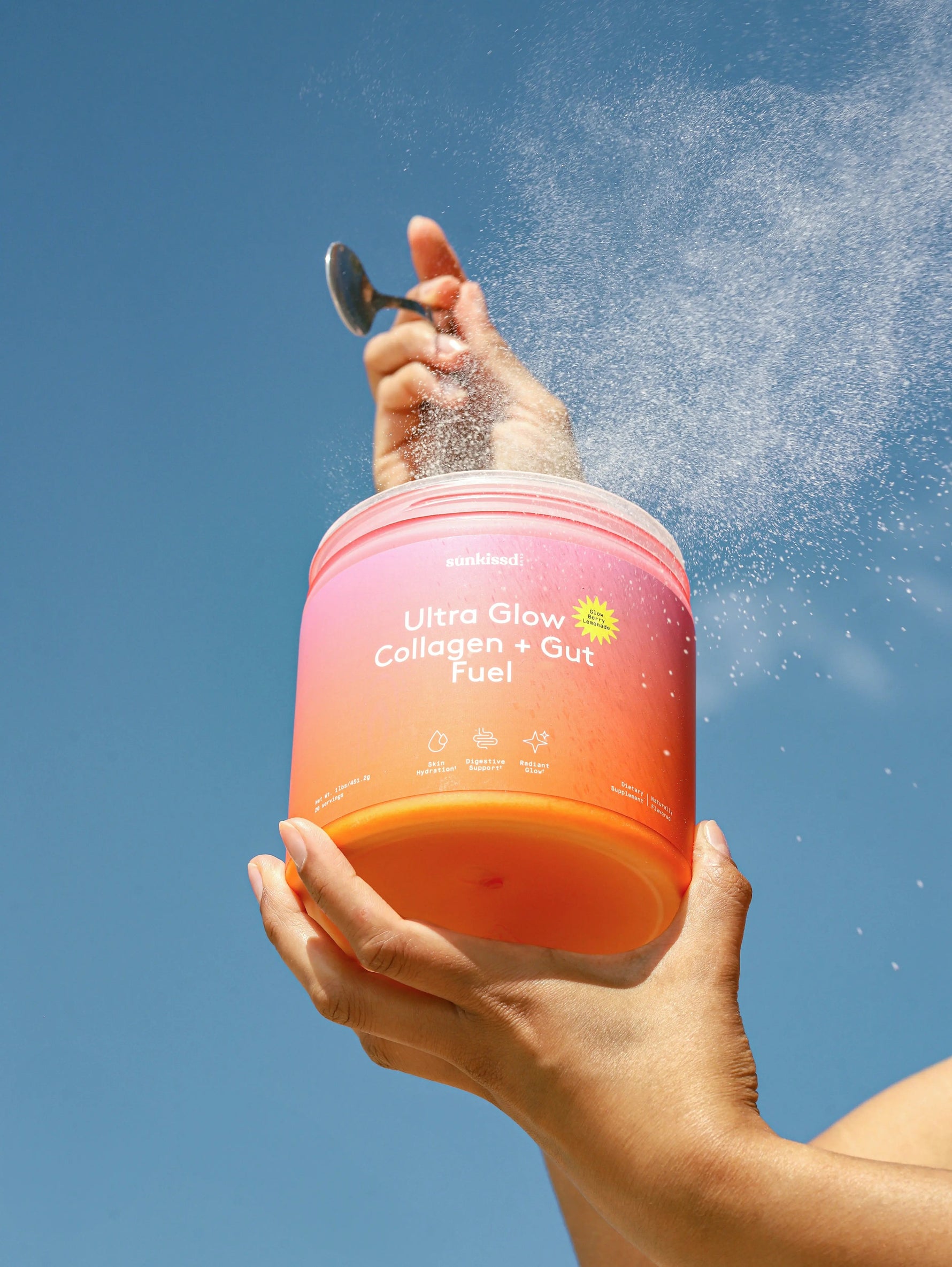 A person holds a pink and orange jar labeled 'umbrahealth Ultra Glow Collagen + Gut Fuel' against a bright blue sky, with powder spraying out of the top and a spoon holding more powder.