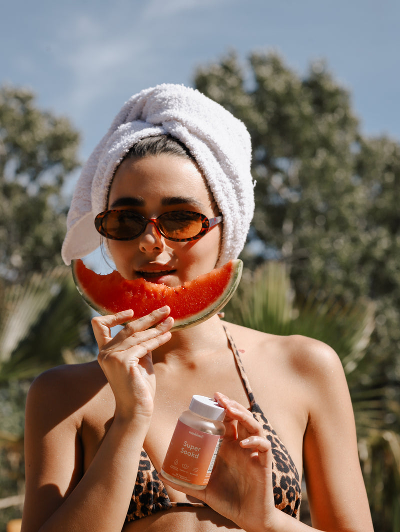 Woman with towel on head holding watermelon and supplement bottle outdoors
